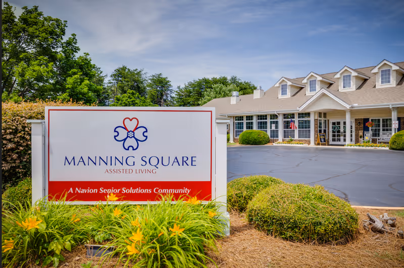 Exterior view of Manning Square Assisted Living facility with a large sign in the foreground displaying the facility name and logo. The building has a beige exterior with multiple windows and dormer windows on the roof. There are bushes, flowers, and a paved driveway in front of the building under a partly cloudy sky.