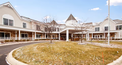 Exterior view of a senior living facility with a circular driveway, a grassy area with some trees, and a large white building with multiple windows and a covered entrance under a clear blue sky.