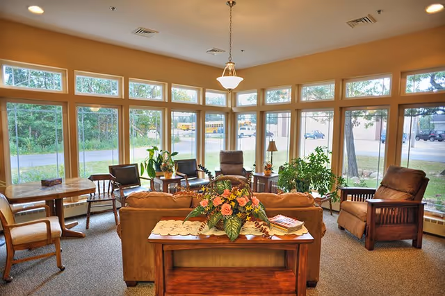 Sunlit senior living lounge with sofas, chairs, plants, and a central flower arrangement near large windows.