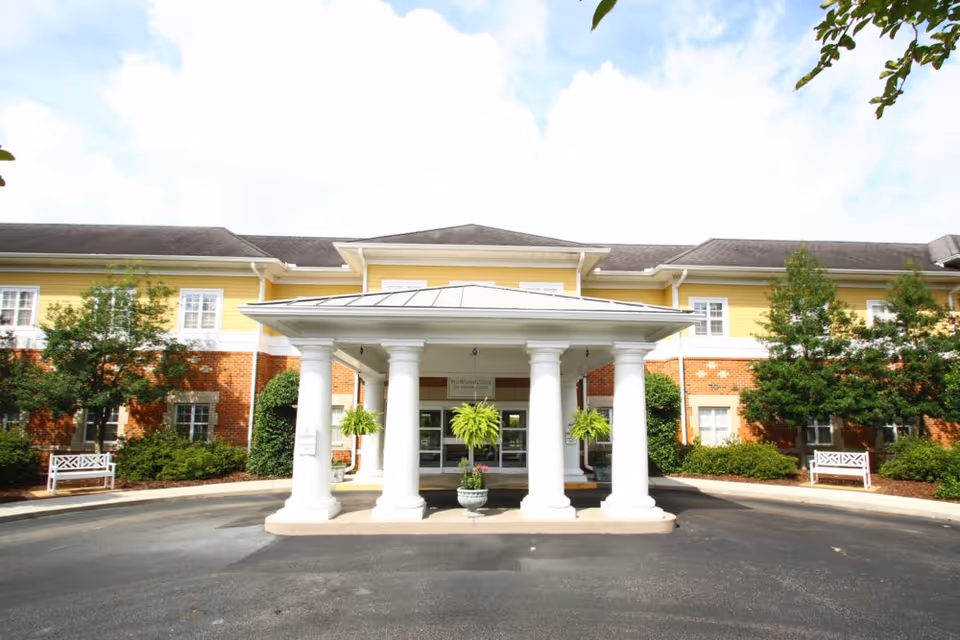 Front exterior view of Morningside of Wilmington facility showing a covered entrance with white columns, potted plants, and a two-story building with yellow and brick facade under a partly cloudy sky.