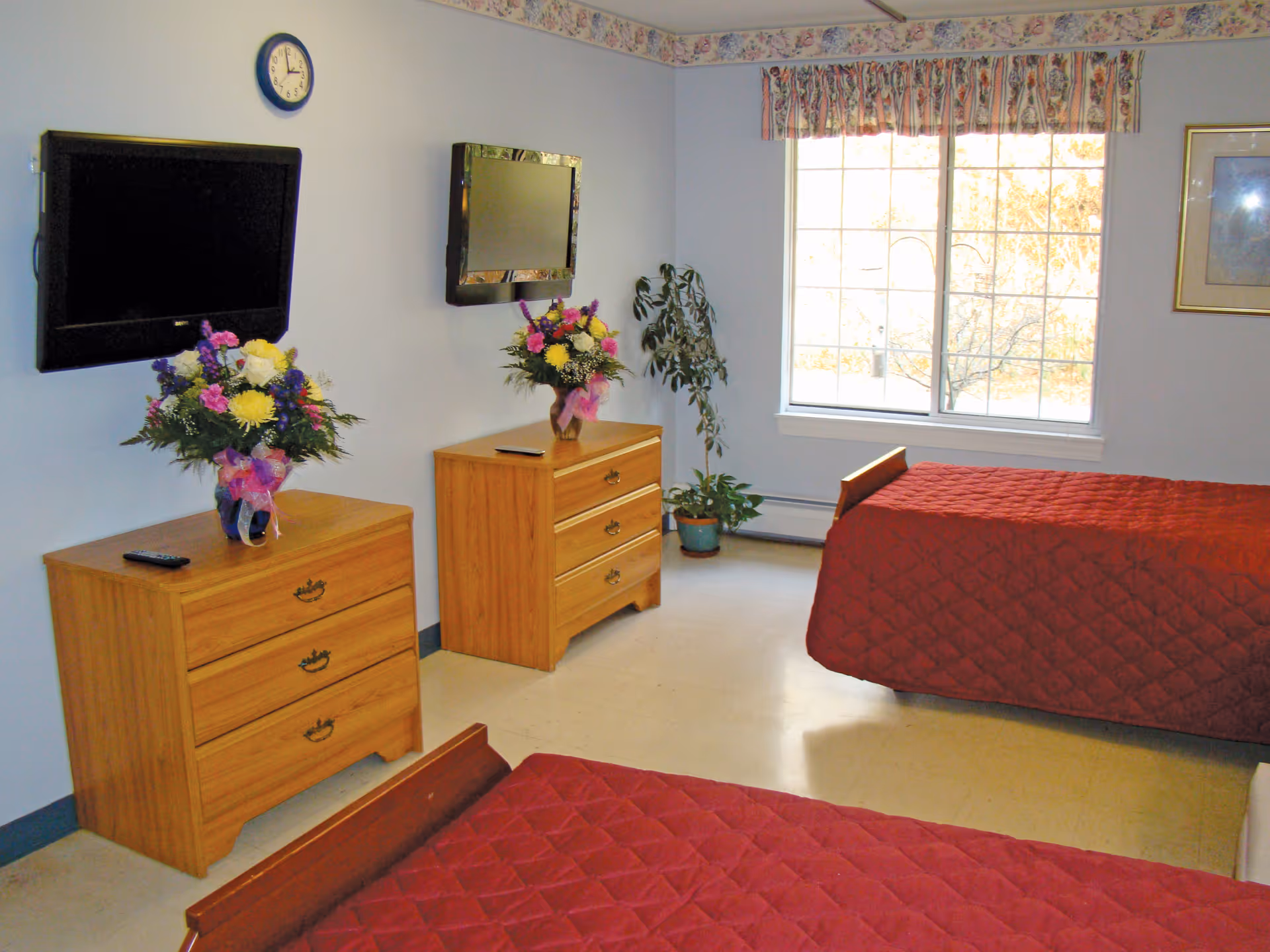 A senior living facility bedroom with two single beds covered in red quilted bedspreads. Each bed has a wooden headboard. There are two wooden dressers, each with a colorful flower arrangement on top and a TV mounted on the wall above. A clock is mounted on the wall above one dresser. A large window lets in natural light, and a potted plant is placed near the window. The walls are light-colored with a floral border near the ceiling.