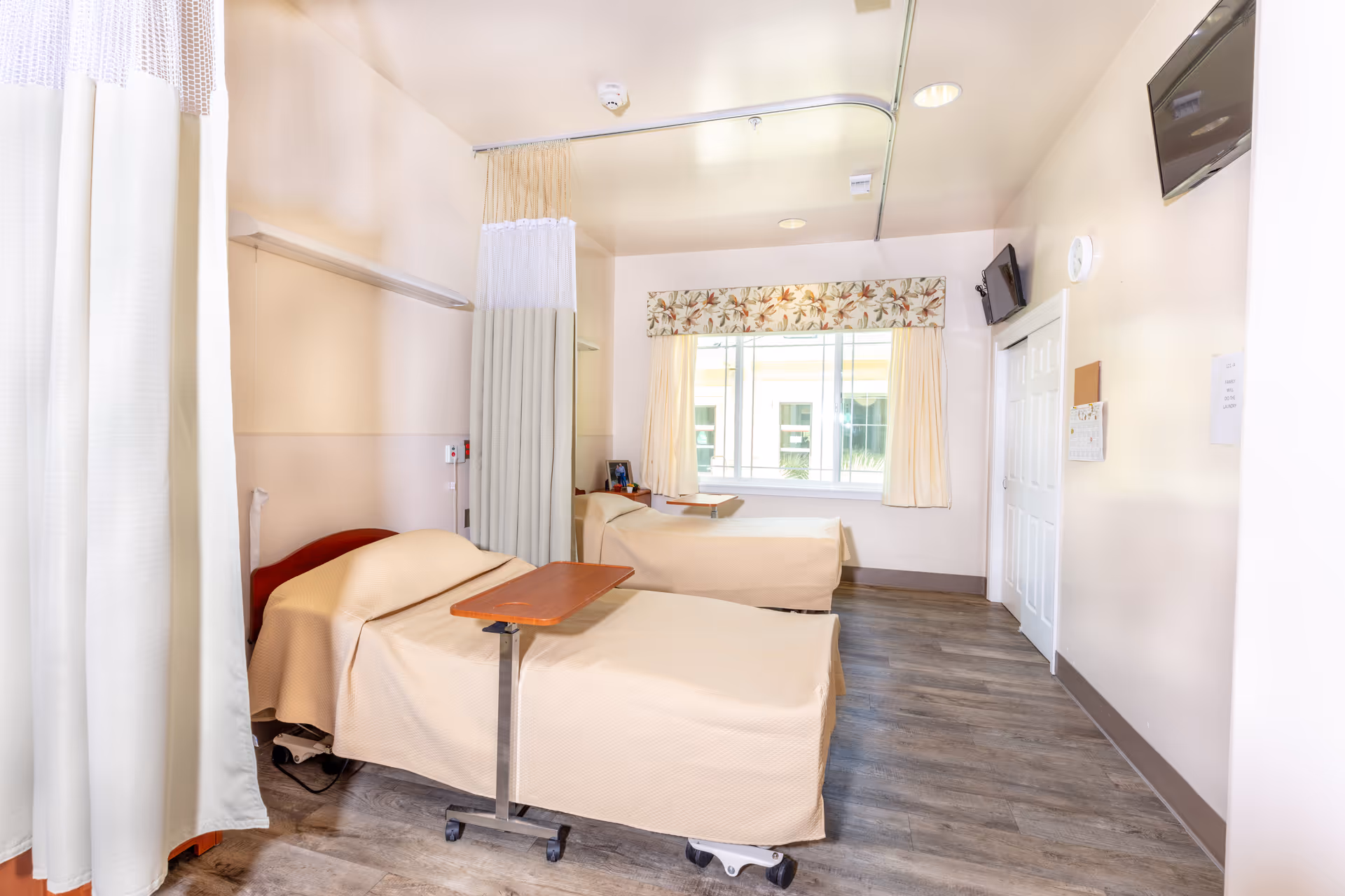 Two hospital beds with privacy curtains in a clean patient room with a window.