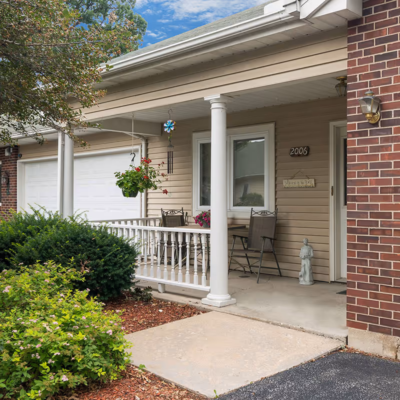 Front porch of a residence with white columns, a small seating area, hanging plants, and house number 2006.