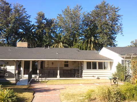 Single-story building with a covered porch and ramp entrance, surrounded by a garden with grass, bushes, and tall trees in the background under a clear blue sky.