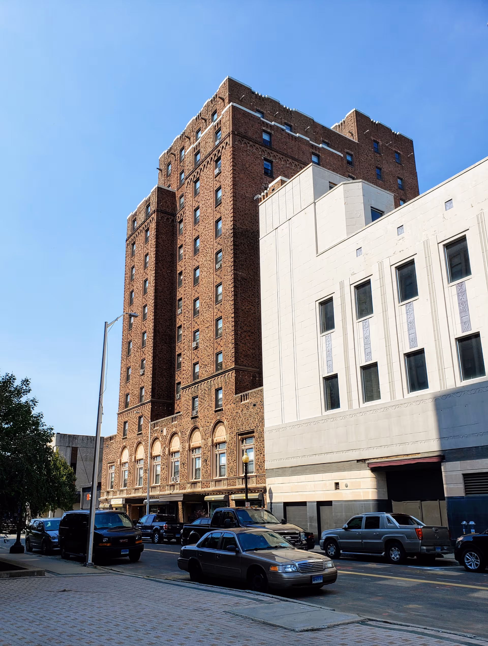 Tall brick senior living building on a city street with parked cars and a white annex
