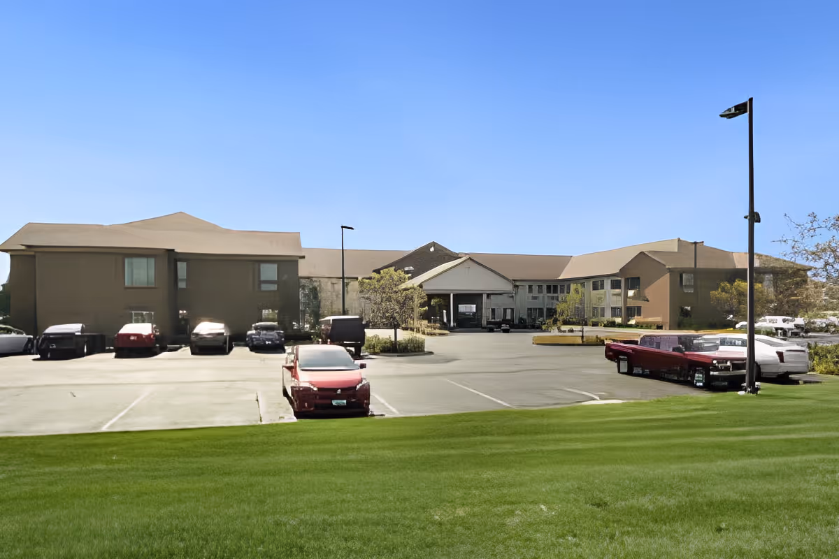 Front view of a senior living facility building with a parking lot, several parked cars, and a green lawn under a clear blue sky.