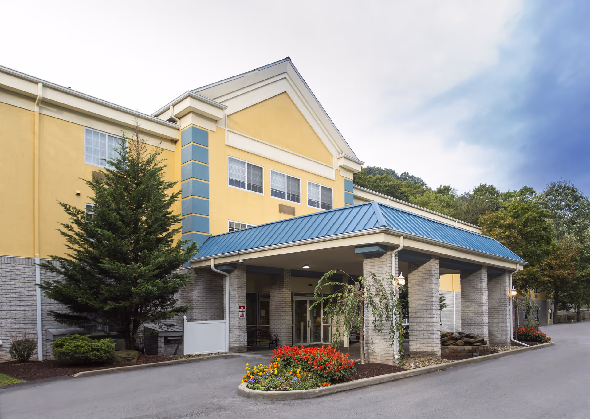 Front entrance of a yellow senior living building with a covered drop-off, blue metal roof, and landscaped flower beds.