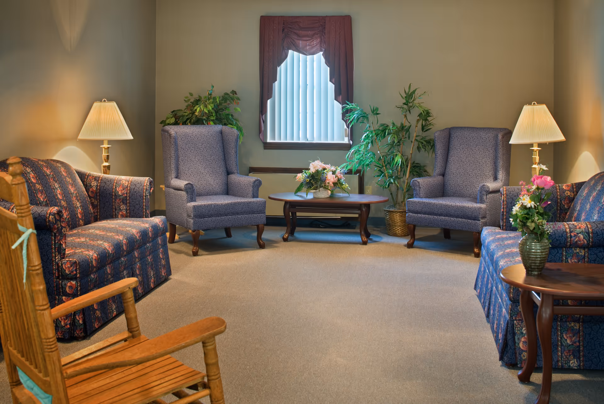 A cozy living room with two patterned blue armchairs, two matching blue sofas, a wooden rocking chair, two table lamps with beige shades, a wooden coffee table with a floral arrangement, and two potted plants near a window with vertical blinds and a purple valance.
