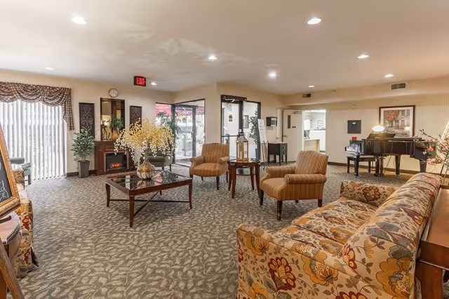 A spacious senior living facility common area with patterned carpet, floral upholstered sofa, two striped armchairs, a glass-top coffee table with a flower arrangement, and a piano in the background. Large windows with vertical blinds and an exit door are visible, along with various decorative plants and framed artwork on the walls.