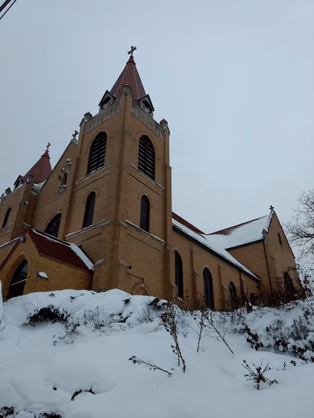 Snow covers a brick church exterior with tall towers against a gray winter sky.