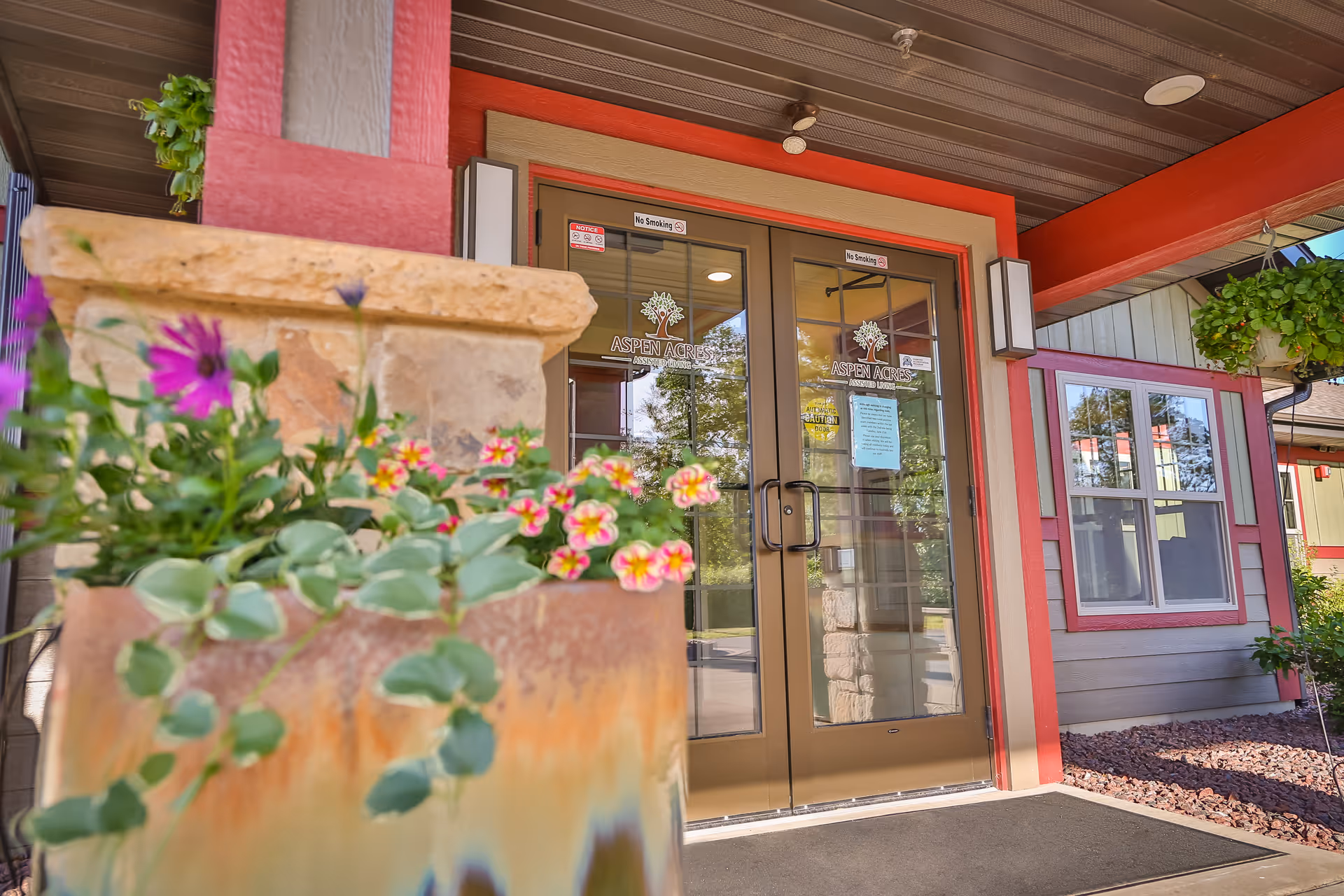 Entrance of Aspen Acres Assisted Living with glass doors, red trim, and potted flowers.