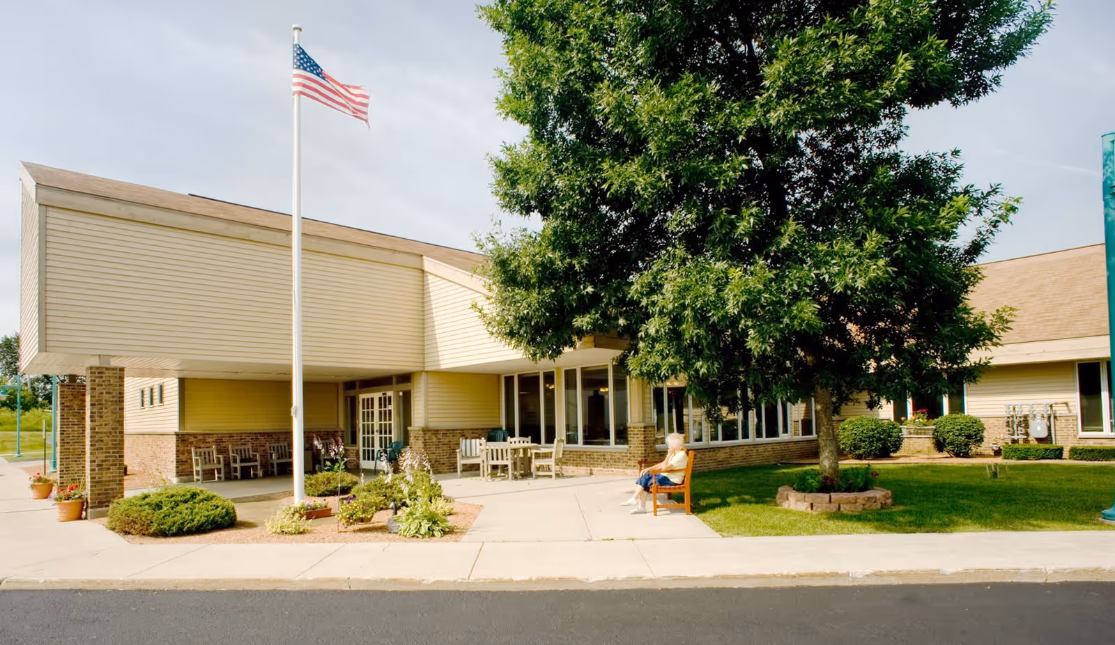 Front entrance of a senior living facility with an American flag on a pole, a covered entry, outdoor seating, a large tree, and a person sitting on a bench.