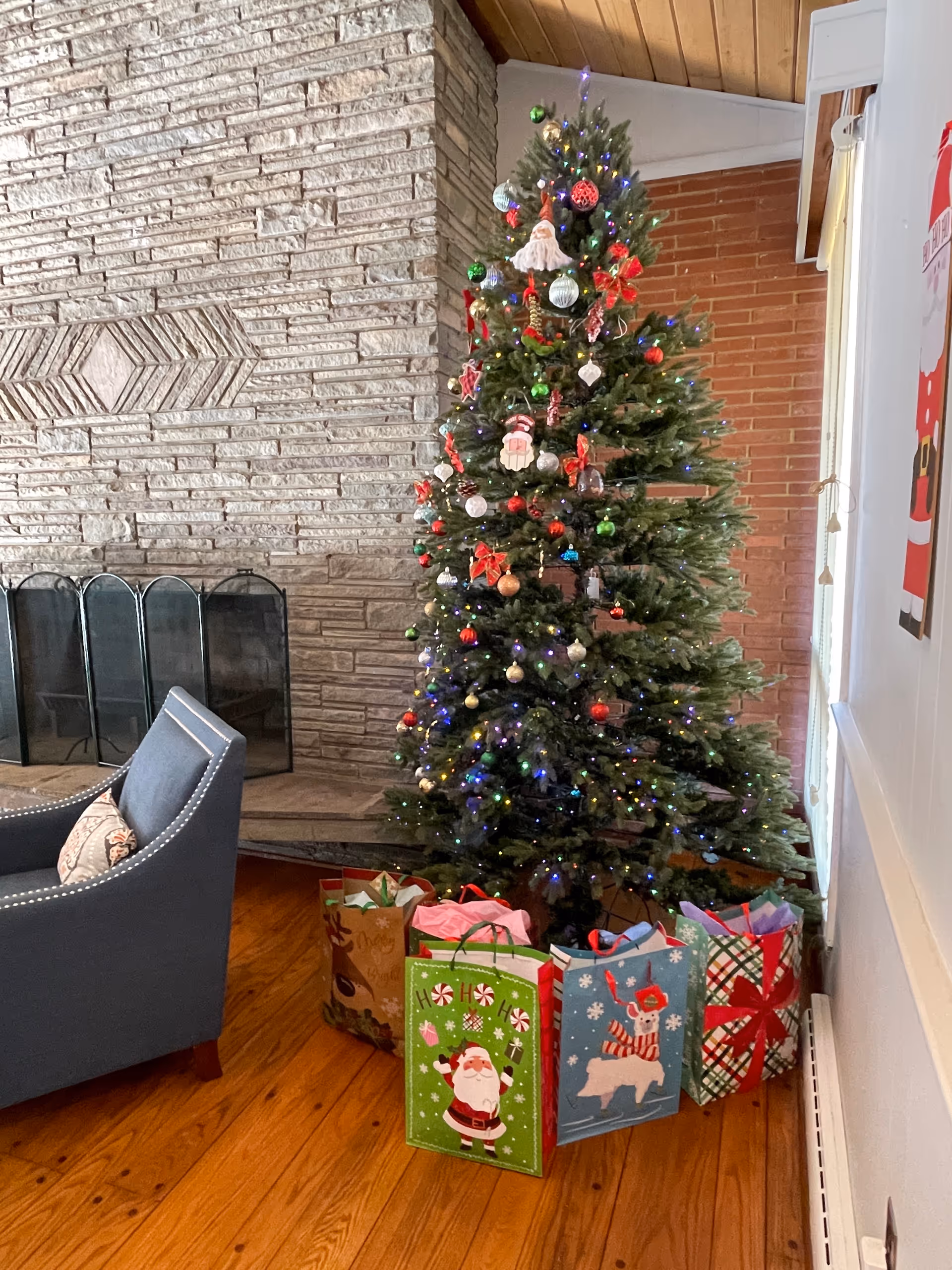 Christmas tree with gift bags beside a stone fireplace in a cozy living area