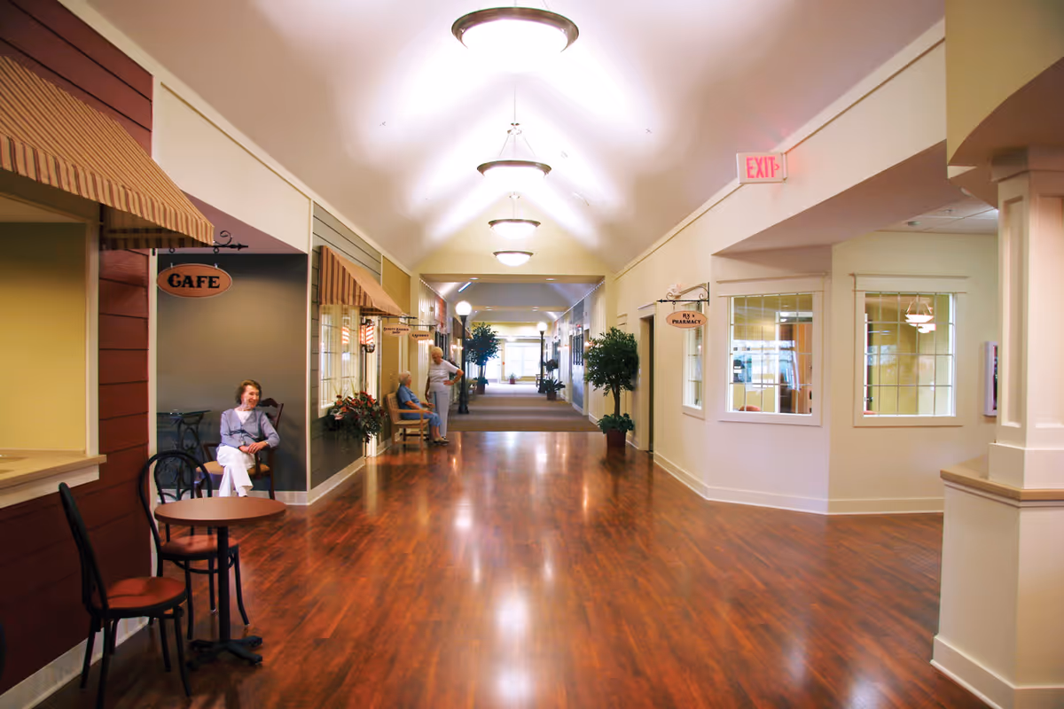 Bright senior living corridor with cafe seating, plants, and warm wood flooring.