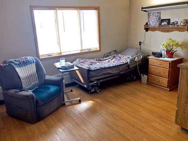A simple, well-lit bedroom with a hospital-style bed covered with a colorful blanket, a wooden nightstand with a potted plant and some items on top, a comfortable armchair with a blanket draped over it, a small adjustable table with a cup and other items, and a window letting in natural light.
