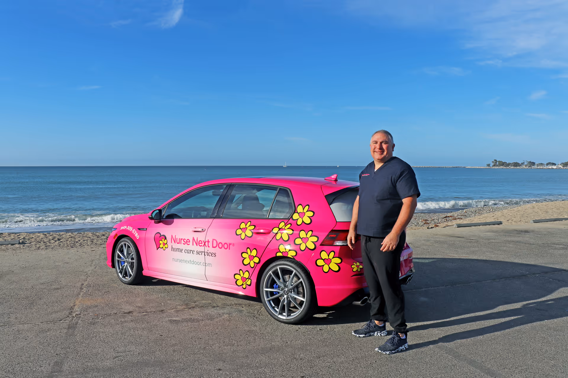 A smiling staff member stands beside a pink floral Nurse Next Door car on a beach.