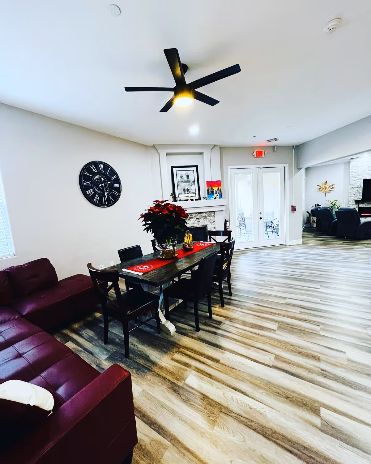 A bright and spacious common area in a senior living facility featuring a dark wooden dining table with six chairs, a red table runner, and a potted plant centerpiece. To the left is a maroon sectional sofa with a white pillow. The room has light wood flooring, a modern black ceiling fan with a light, a large wall clock, and a fireplace with framed artwork above it. Double glass doors lead to an outdoor patio area with seating. In the background, there are additional seating areas with black recliners and decorative plants.