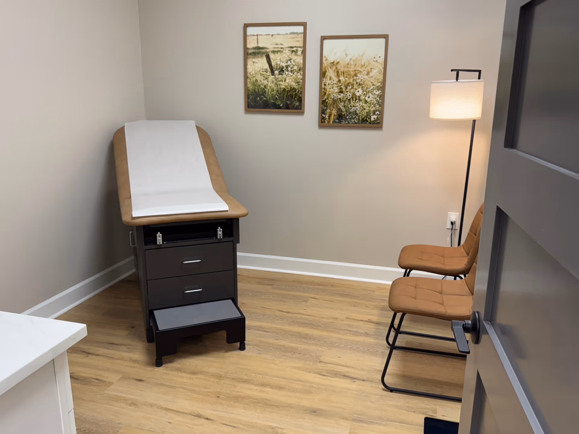 Exam room with a medical chair, drawers, two brown chairs, and wall art.