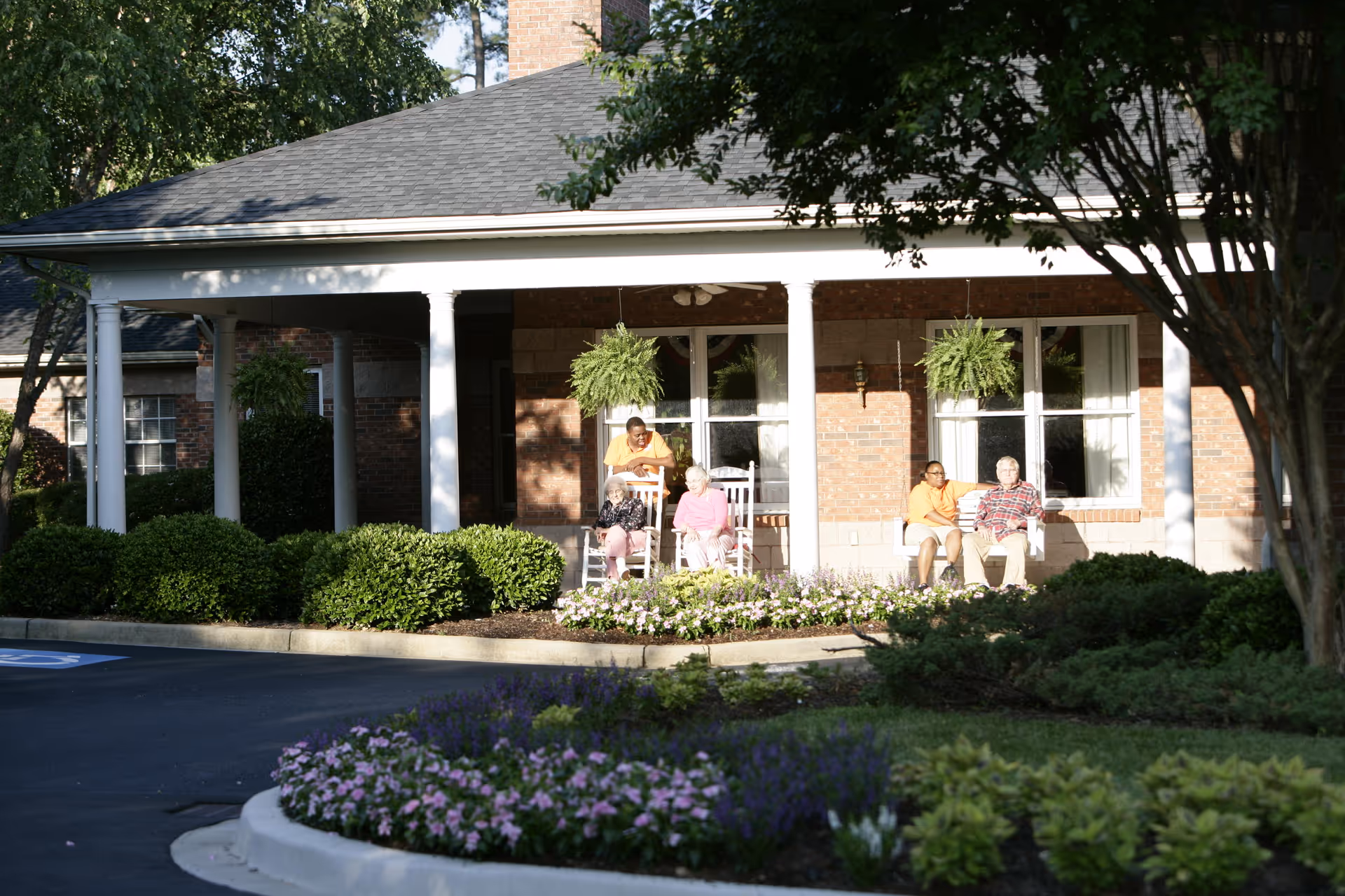 Four elderly individuals sitting and relaxing on rocking chairs and a bench on the covered front porch of a brick building surrounded by greenery and flower beds.