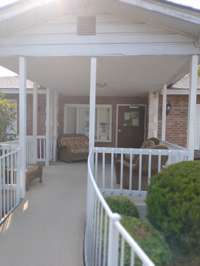 Covered front entrance of a brick assisted-living building with a ramp, white railings and wicker seating on the porch.
