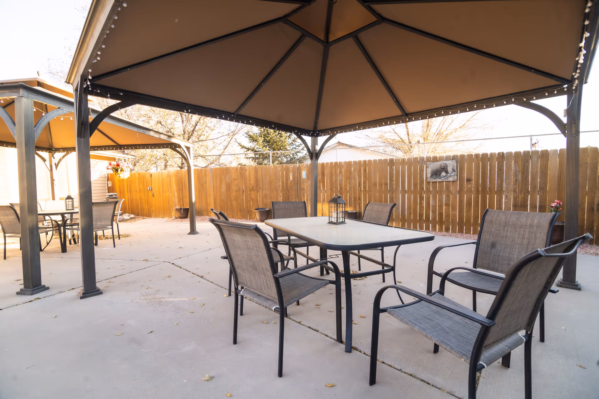 Outdoor patio area with two covered gazebos, each containing a table and several chairs. The area is enclosed by a wooden fence and decorated with a small wall hanging and potted plants.