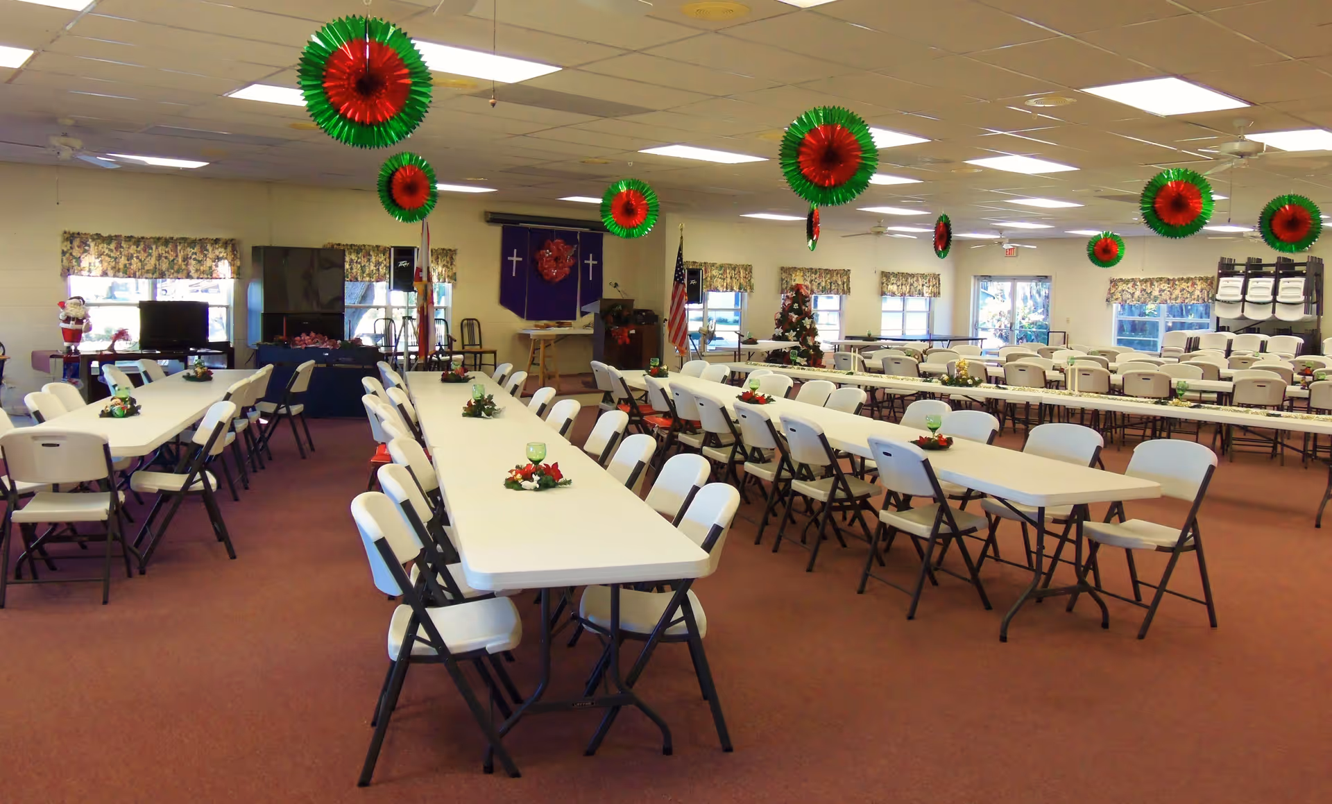 A large dining room with multiple long tables and white folding chairs arranged neatly. The tables have small holiday-themed centerpieces. Red and green circular decorations hang from the ceiling. Windows with floral curtains line the walls, and an American flag and a decorated Christmas tree are visible in the background.