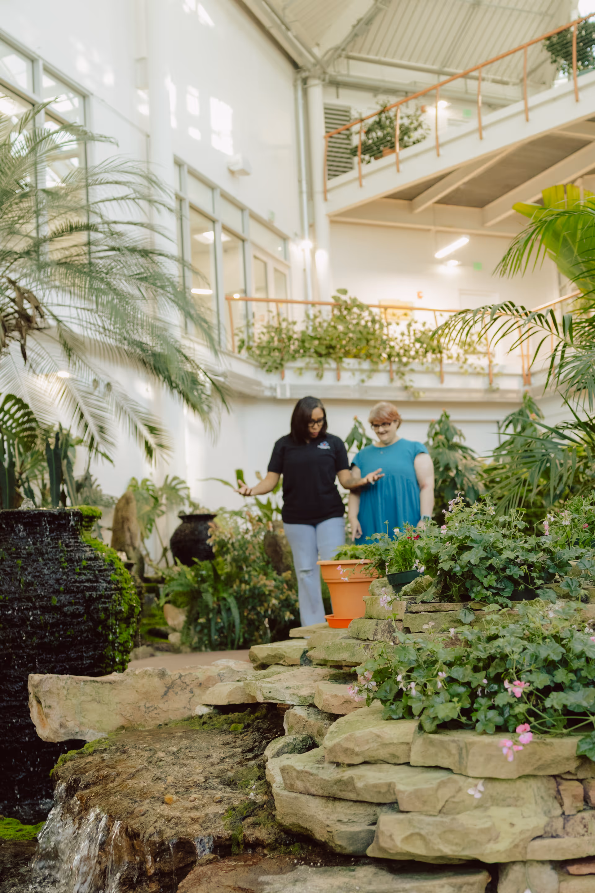 Two women walk through a lush indoor garden atrium with rocks and a water feature.