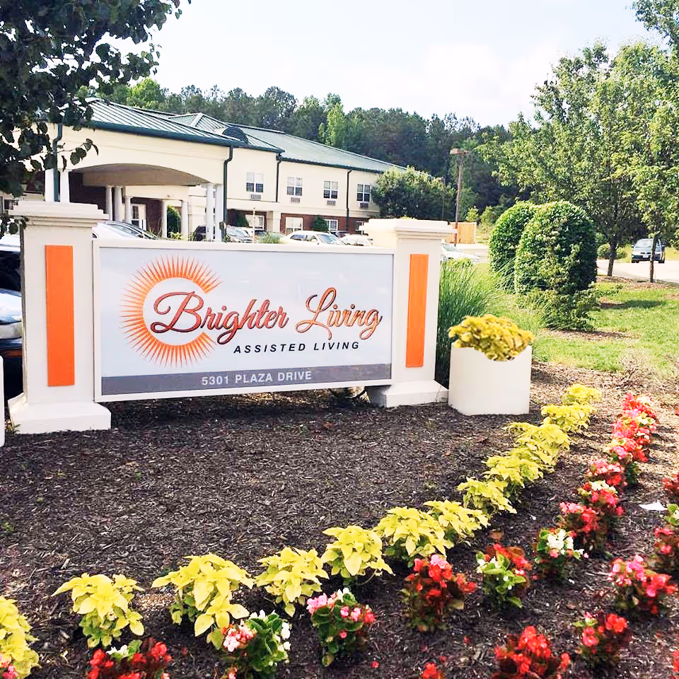 Outdoor view of the entrance sign for Brighter Living Assisted Living facility, surrounded by landscaped flowers and greenery, with the building and parking area visible in the background.