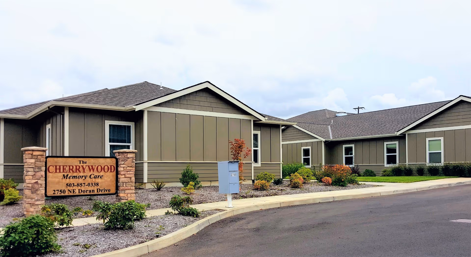 Exterior view of Cherrywood Memory Care facility showing a single-story building with gray siding and a shingled roof. There is a landscaped area with small shrubs and plants in front of the building, and a sign that reads 'The Cherrywood Memory Care 503-857-0338 2750 NE Doran Drive'. The sky is cloudy.