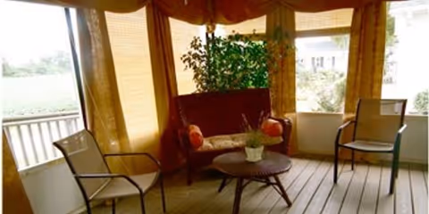 Enclosed porch/sunroom with a wicker loveseat, two chairs, a round table and a potted plant in front of windows with blinds.