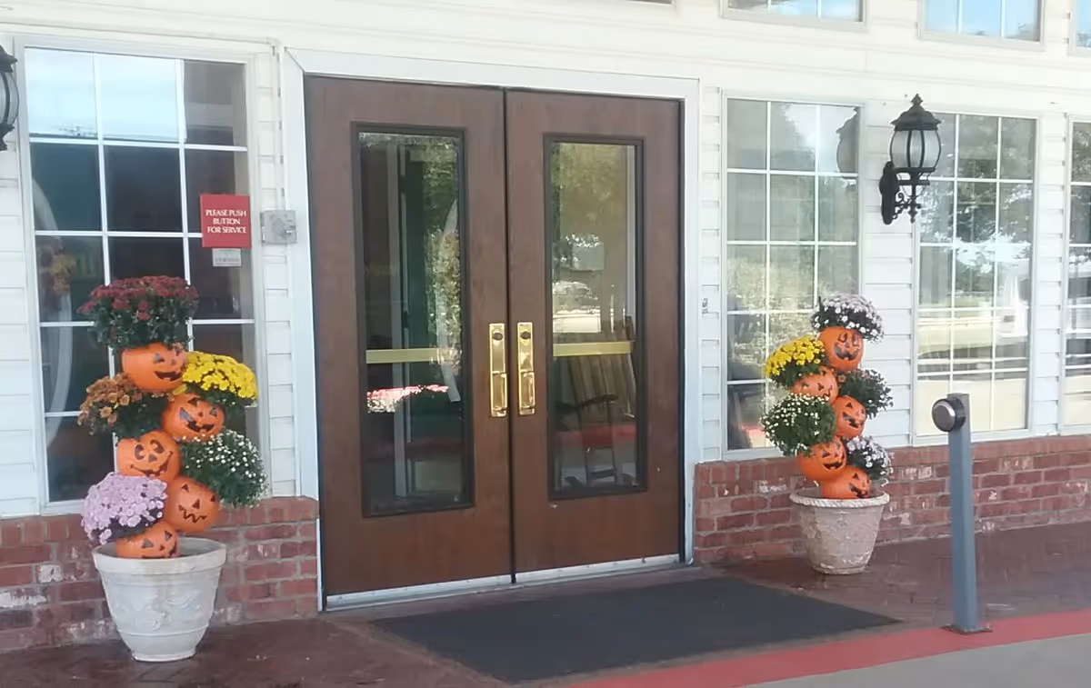 Entrance to a building with double wooden doors featuring glass panels. On either side of the doors are large windows and decorative flower pots with stacked orange pumpkin faces and colorful flowers. A black lantern-style light fixture is mounted on the wall to the right of the doors.