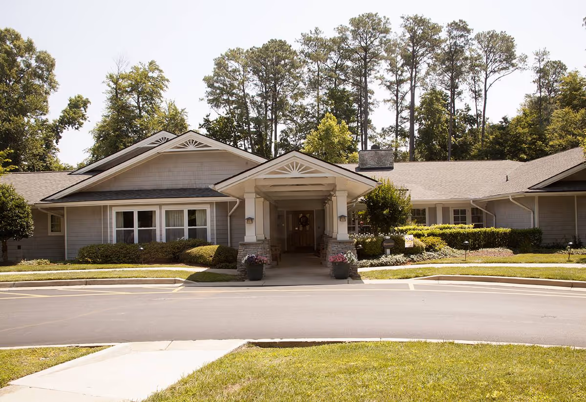 Front exterior of a single-story senior living building with a covered entrance, potted flowers, and surrounding landscaping.