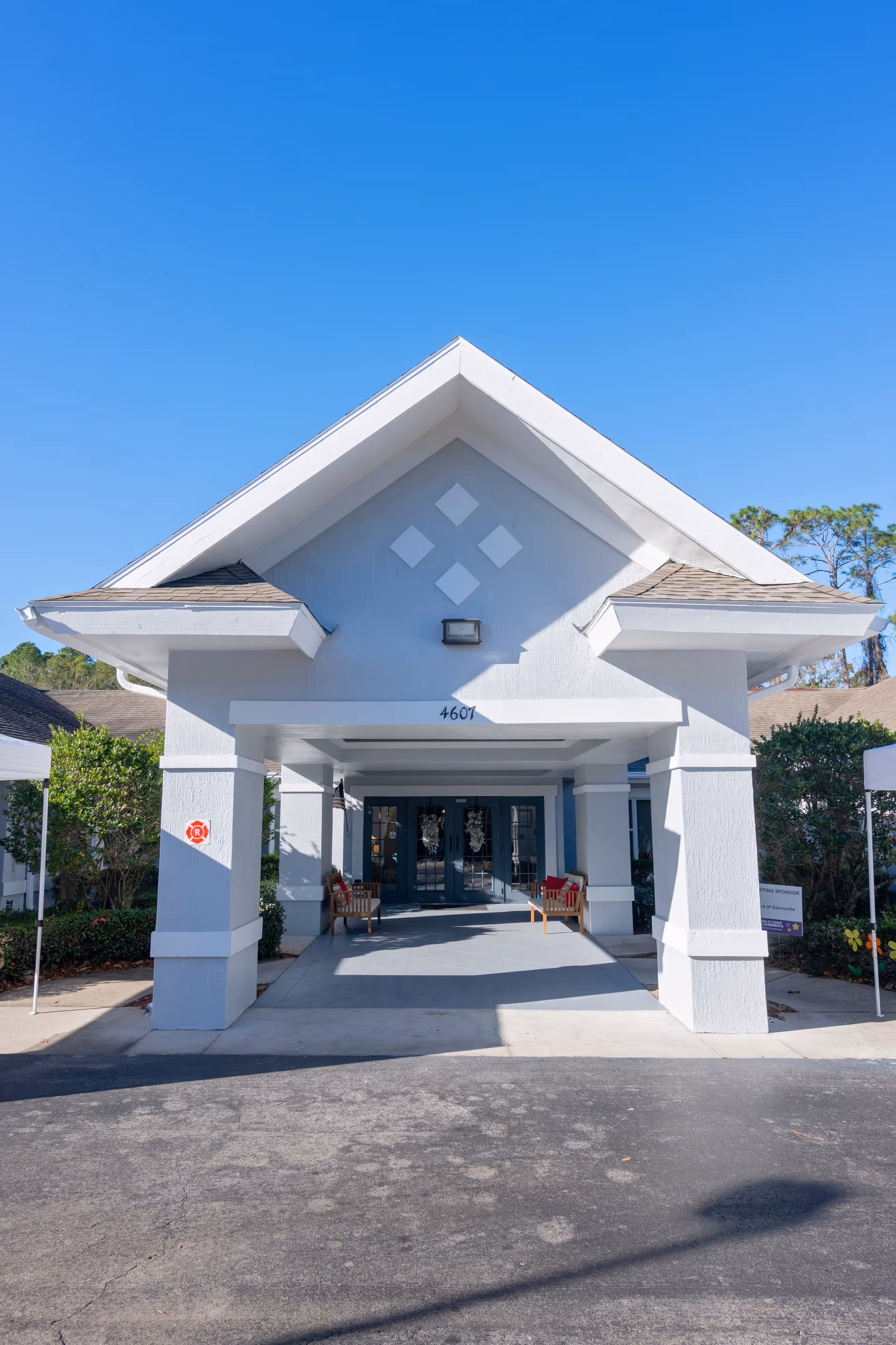 Front entrance of a building with a covered driveway, white pillars, and a peaked roof under a clear blue sky. There are benches with red cushions on either side of the entrance and greenery around the building.