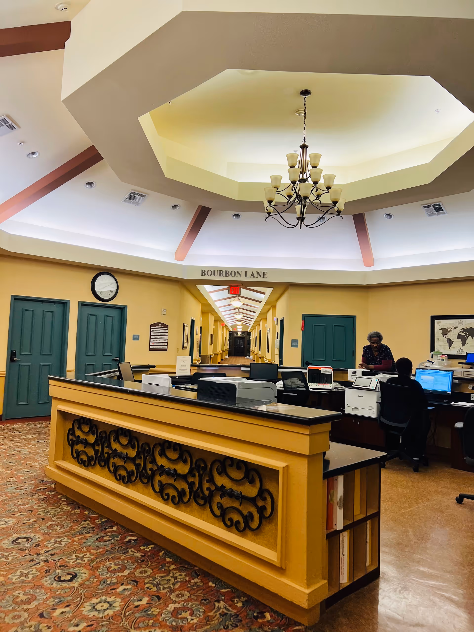 Reception desk in a warm senior living lobby, with a long hallway labeled Bourbon Lane.