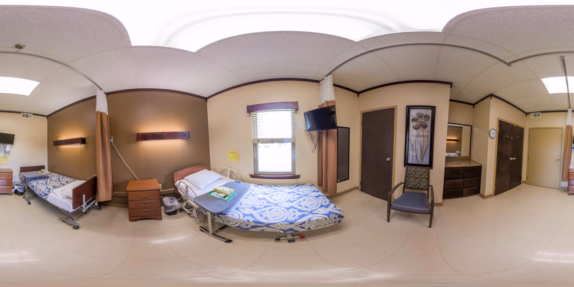 Shared nursing facility patient room with two adjustable beds, a nightstand, wall-mounted TV, chair, and a window.