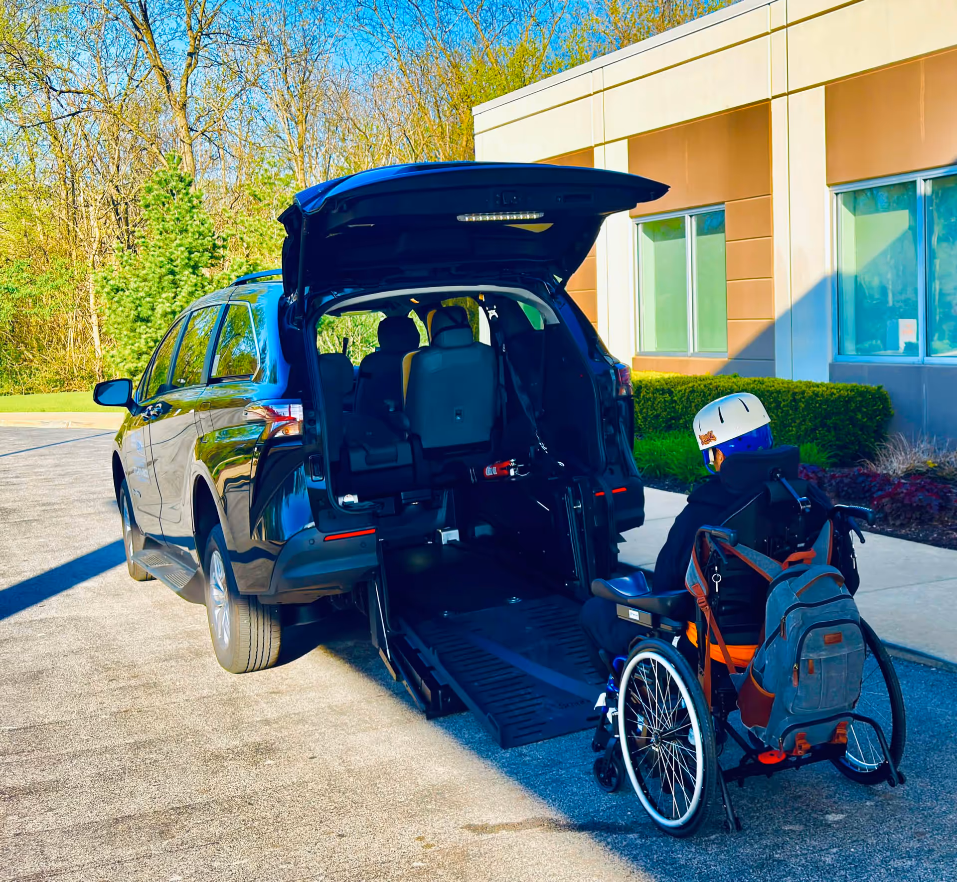 Wheelchair user beside a van with a ramp at a modern building exterior.