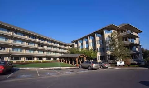A multi-story senior living facility exterior with a front entrance canopy and parked cars.