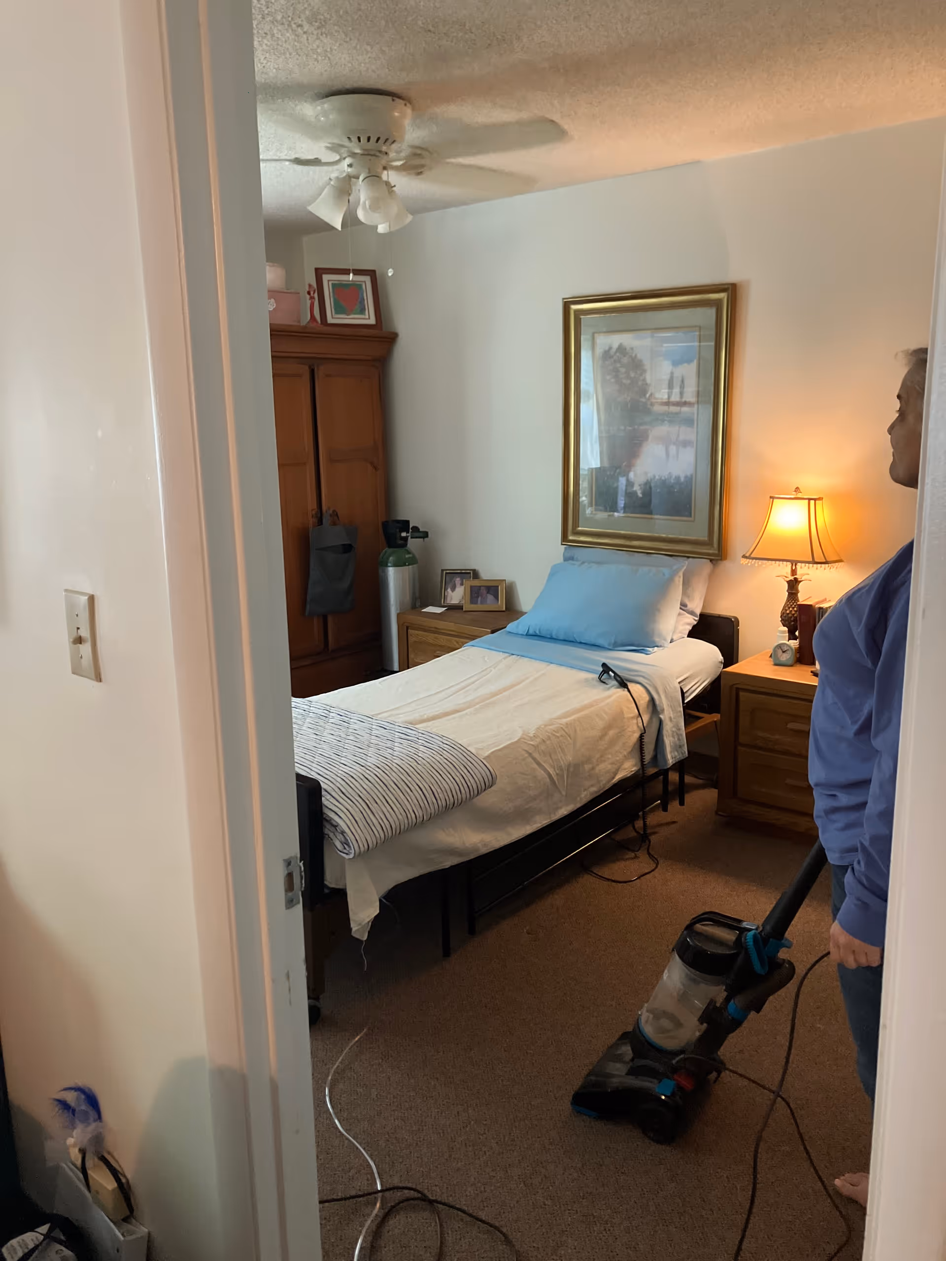A small bedroom in a senior living room with a bed, furnishings, and vacuum.