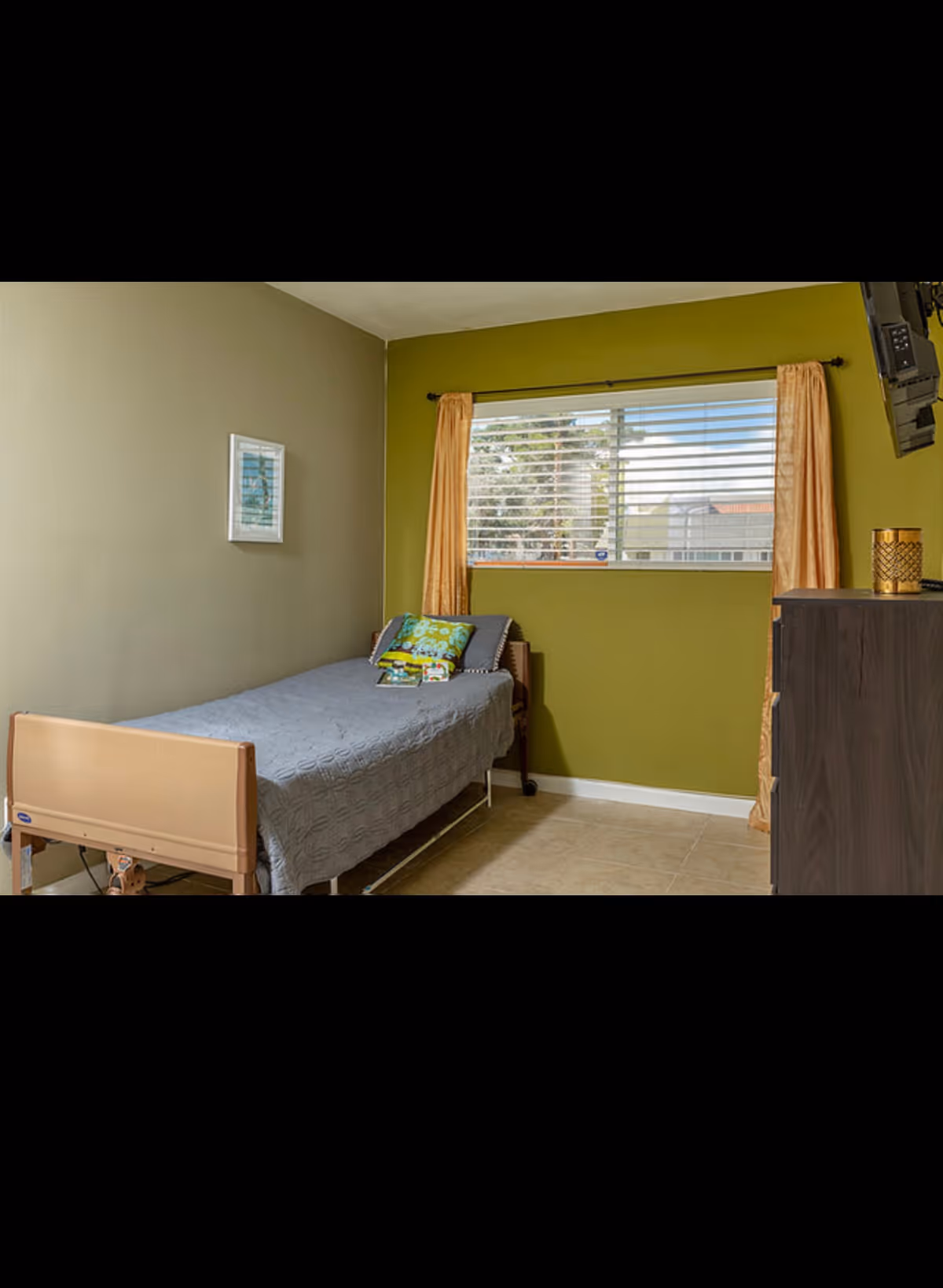 Small single bedroom with an adjustable bed against a green wall, a window with curtains, and a dresser with a wall-mounted TV.