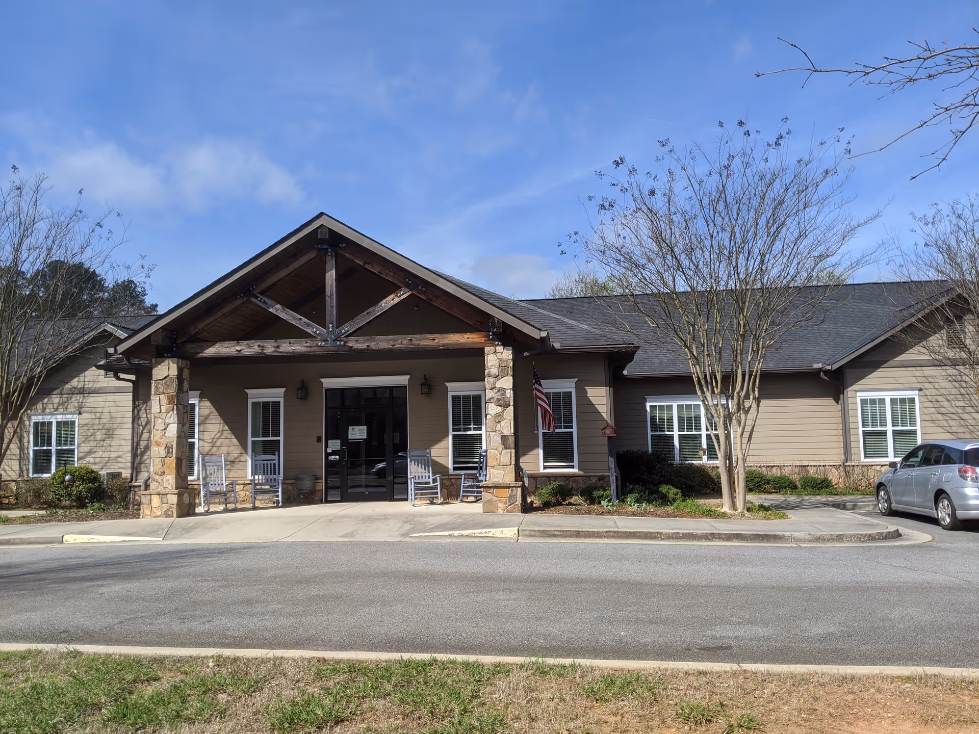 Front view of Heritage Hospice building with stone entrance, rocking chairs, American flag, and blue sky.