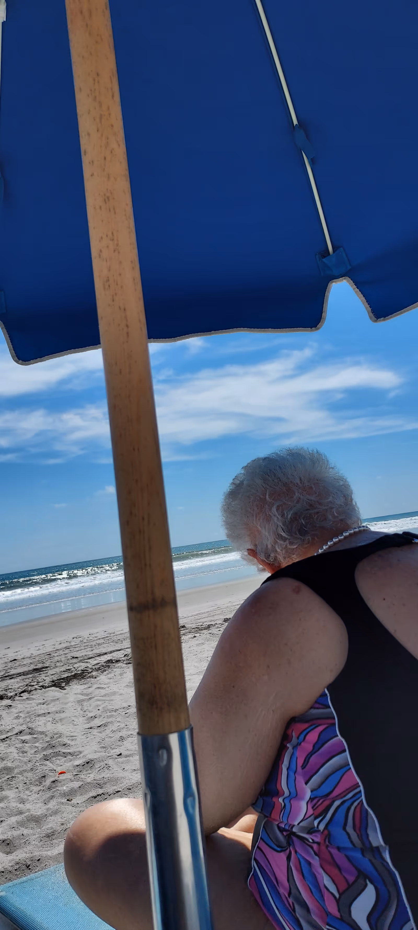 A person sits under a blue beach umbrella on a sandy shore with the ocean behind.