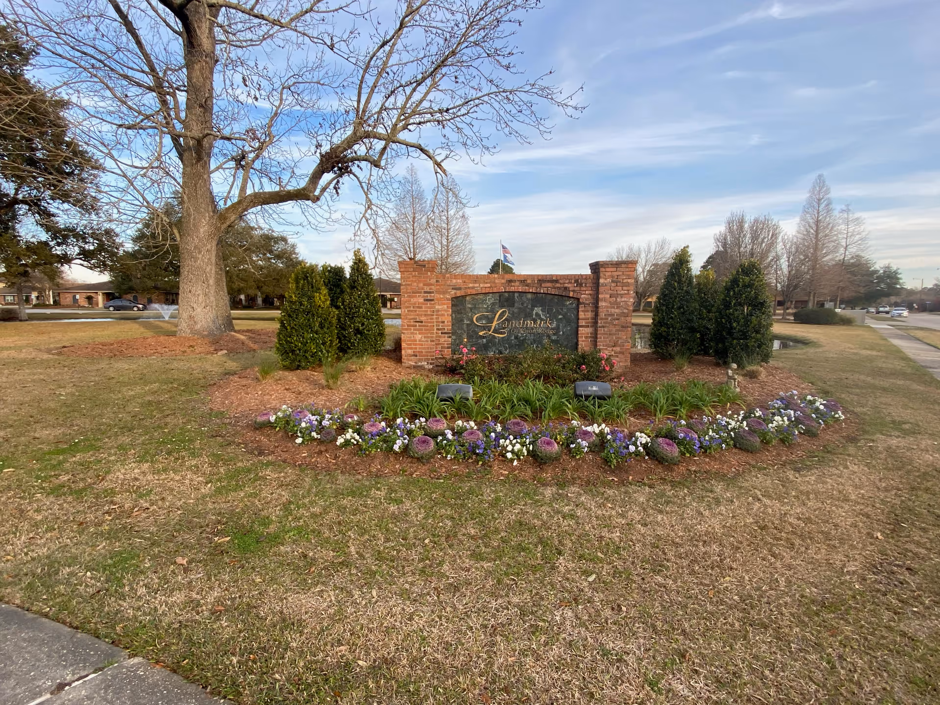 A landscaped outdoor area featuring a brick sign with the text 'Landmark of Baton Rouge' surrounded by green shrubs and a flower bed with purple and white flowers. There are trees and a sidewalk visible in the background under a partly cloudy sky.