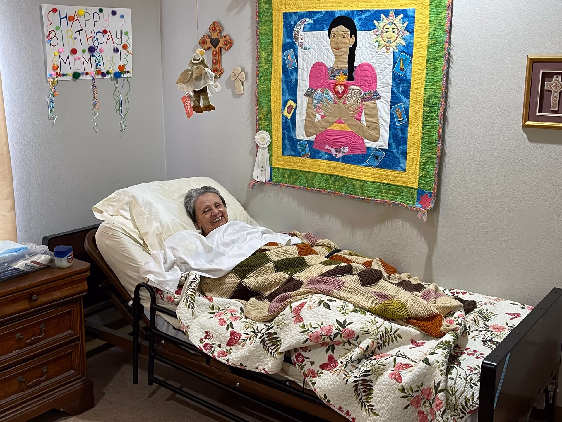 An older woman smiling while lying in a decorated personal-care bedroom bed covered with quilts and wall hangings on the walls.