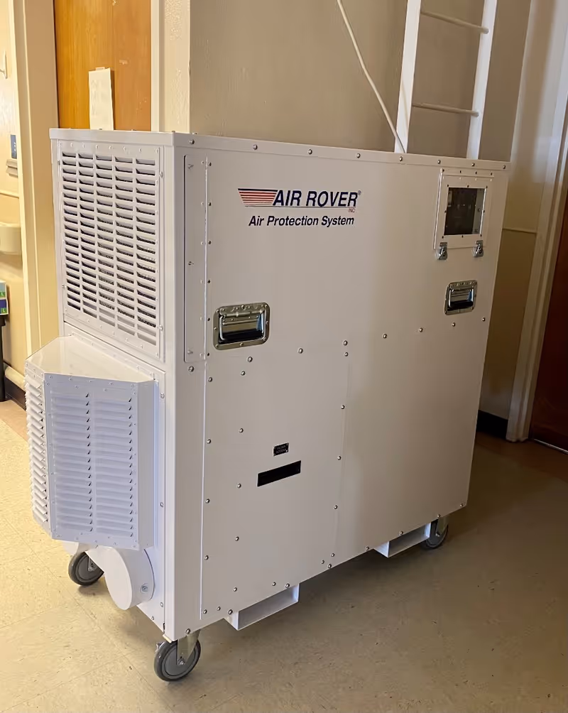 A large white portable air filtration unit labeled 'AIR ROVER Air Protection System' on wheels, placed indoors on a tiled floor near a wooden door and a beige wall.