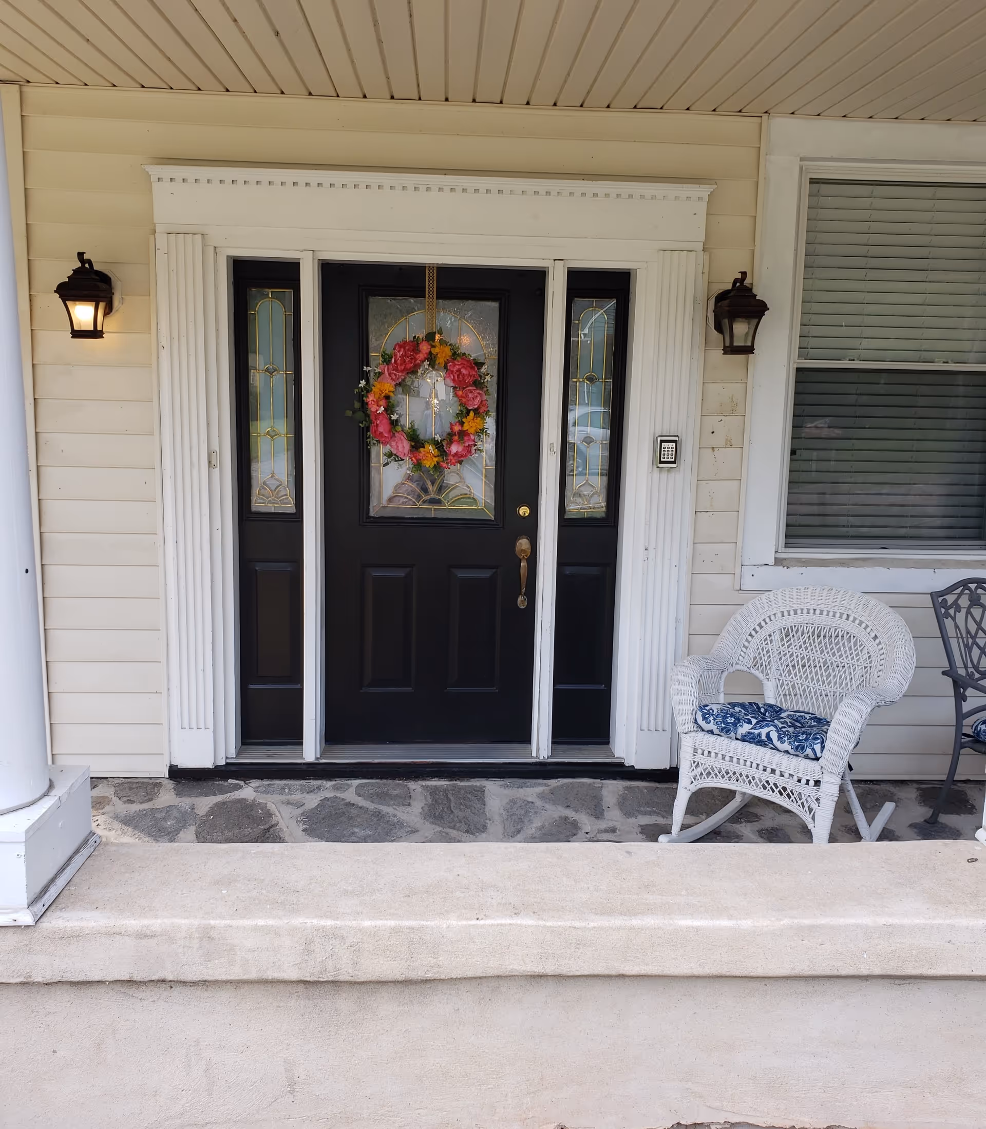 Front entrance of a house with a black door, wreath, lanterns, and wicker chair.