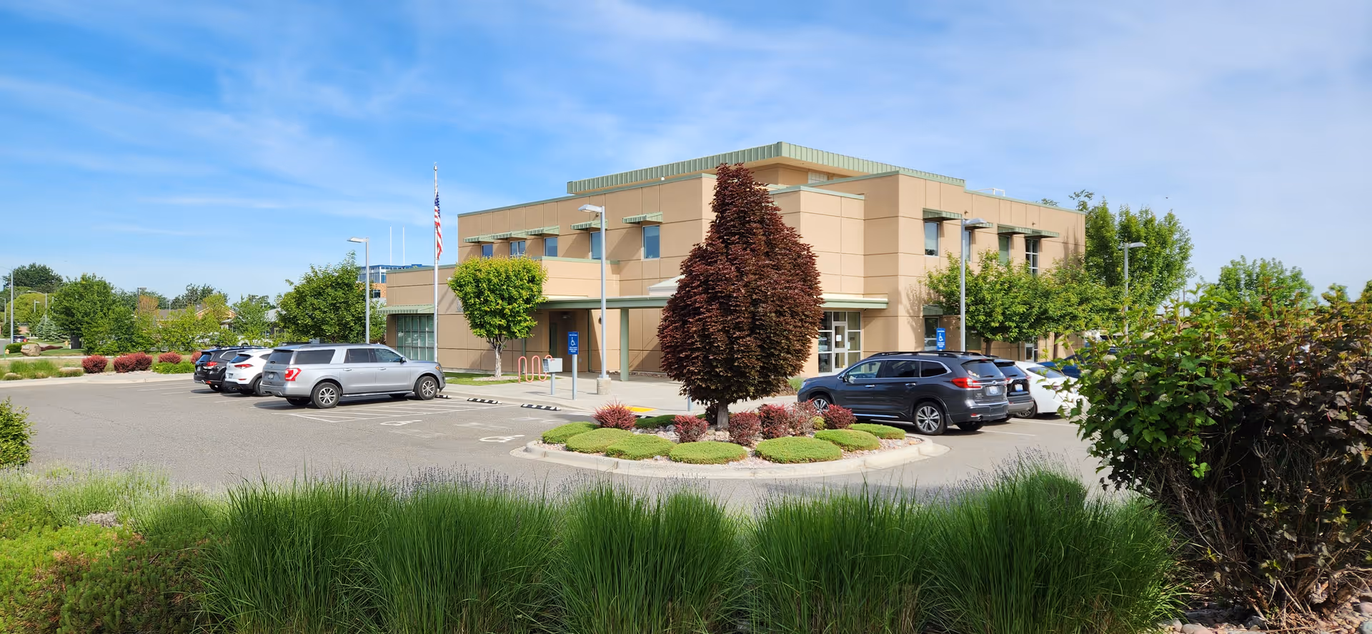Exterior view of a tan senior living facility with a circular landscaped island and parked cars.