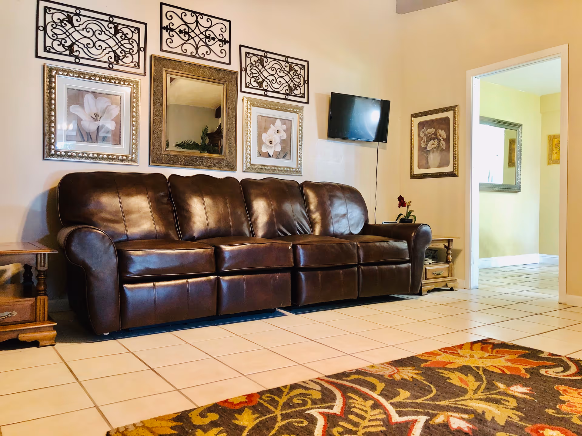 A living room with a dark brown leather sofa against a beige wall. Above the sofa are three framed floral pictures and a decorative mirror with ornate metal wall art. A small flat-screen TV is mounted on the wall to the right. There are two wooden side tables on either side of the sofa, one with a small plant. The floor is tiled with light-colored tiles, and a colorful patterned rug is partially visible in the foreground. An open doorway leads to another room with yellow walls and a mirror on the wall.