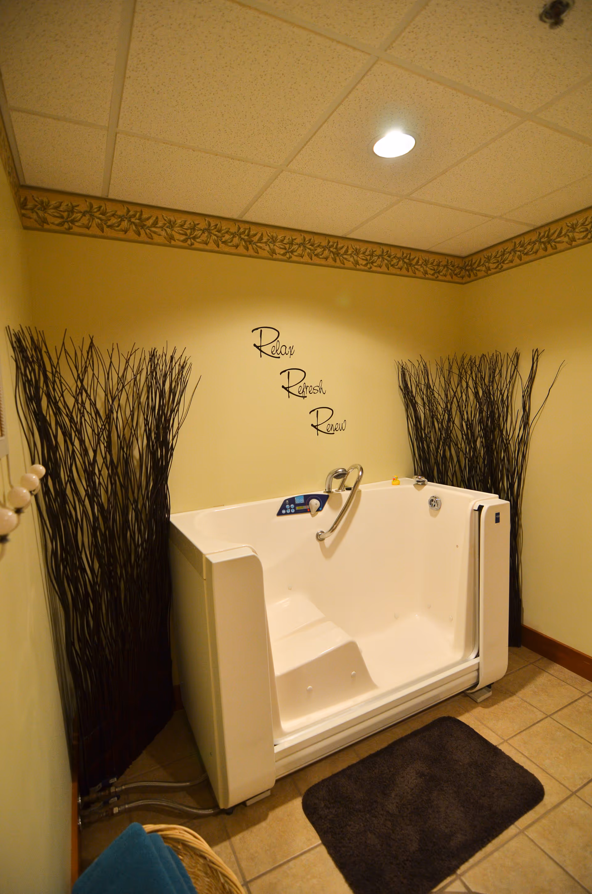 A walk-in bathtub in a bathroom with beige walls and tiled floor. The bathtub has a built-in seat and safety handle. On the wall above the tub, there are decorative words that say 'Relax Refresh Renew'. Two tall bundles of decorative twigs are placed in the corners behind the tub. A dark bath mat is on the floor in front of the tub.