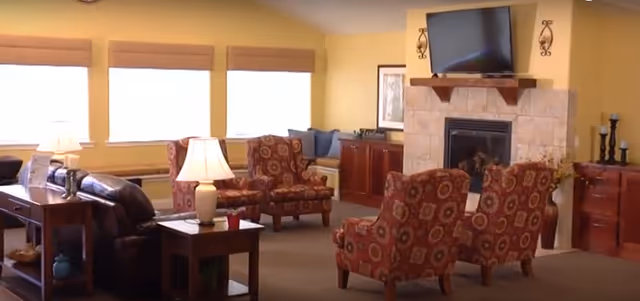 Cozy lounge area with chairs, sofa, fireplace, and TV in a senior living facility.