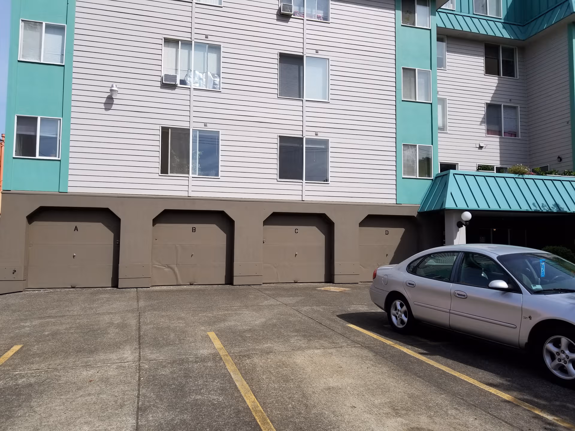 Multi-story building with turquoise accents, row of garage doors labeled A-D, and a parked silver car.